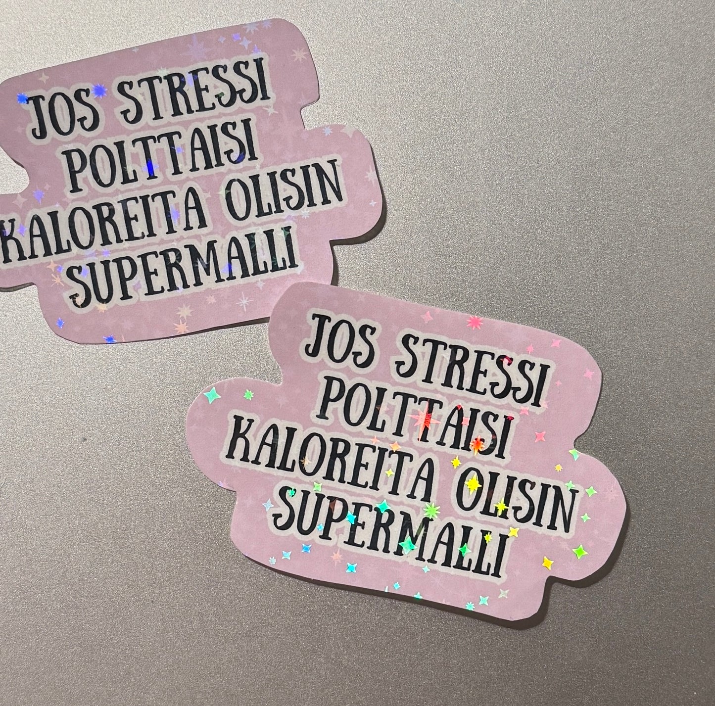 Sateenkaaren väreissä kimaltava&nbsp; laminoitu hologrammitarra, jossa teksti Jos stressi polttaisi kaloreita olisin supermalli – kotimaista käsityötä