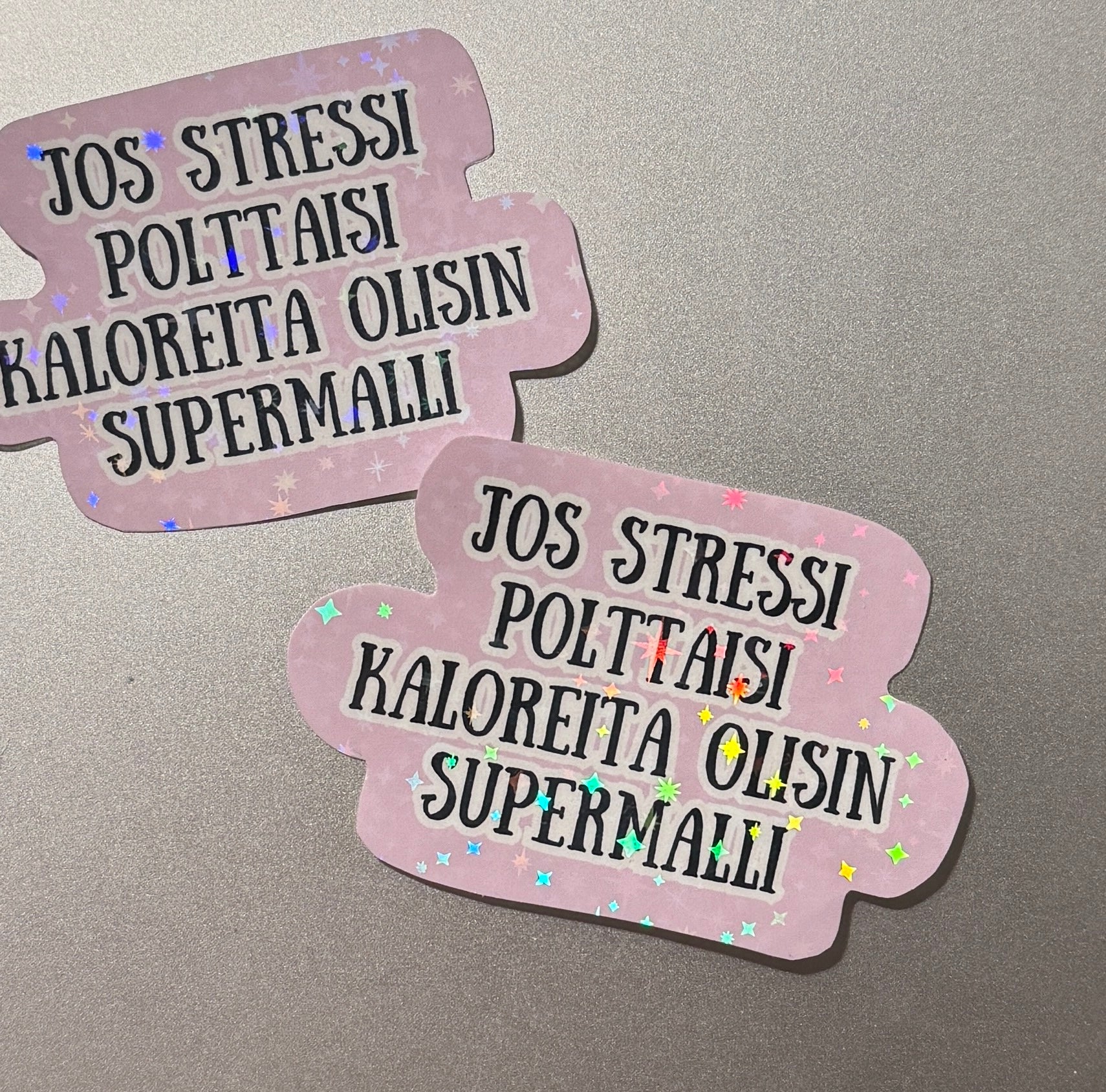 Sateenkaaren väreissä kimaltava&nbsp; laminoitu hologrammitarra, jossa teksti Jos stressi polttaisi kaloreita olisin supermalli – kotimaista käsityötä